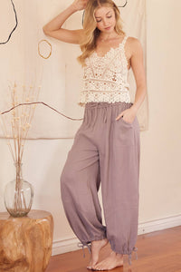 Woman wearing a white lace top and purple pants standing in a room with a wooden table and decorative items.