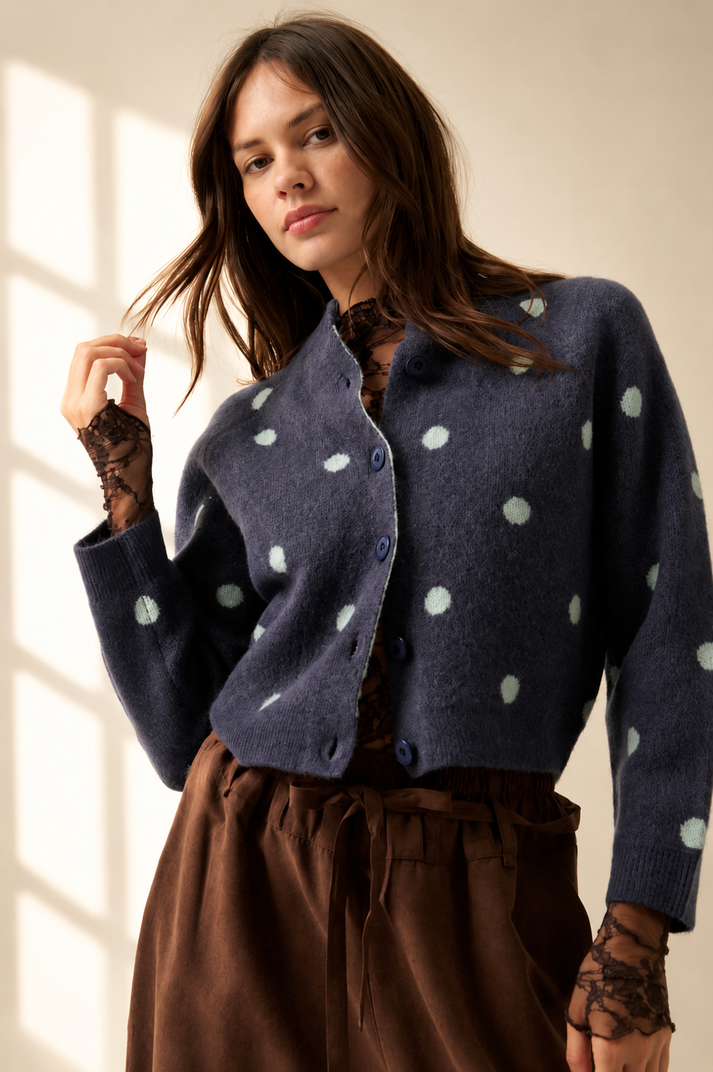 A woman wearing a navy blue fuzzy cropped cardigan with white polka dots, styled over a brown lace long-sleeve top and a brown skirt, standing in a softly lit room.