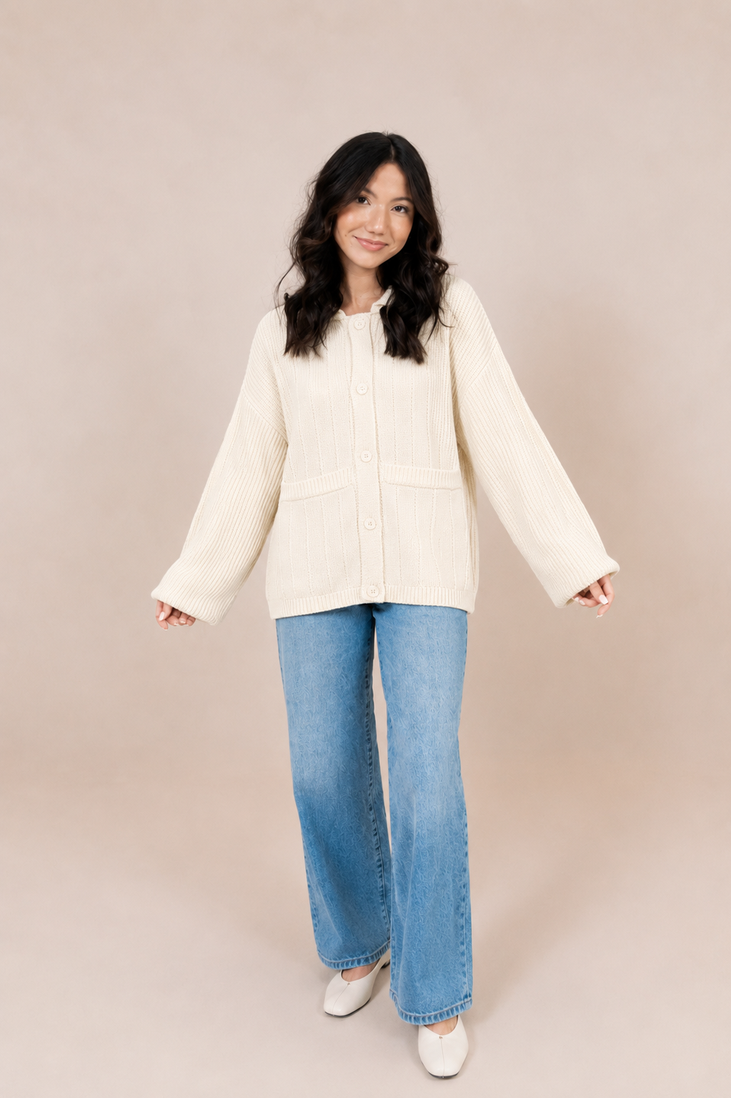 A woman modeling a cream-colored, long-sleeve button-up jacket with front patch pockets, paired with light blue wide-leg jeans and white shoes, against a solid beige studio background.