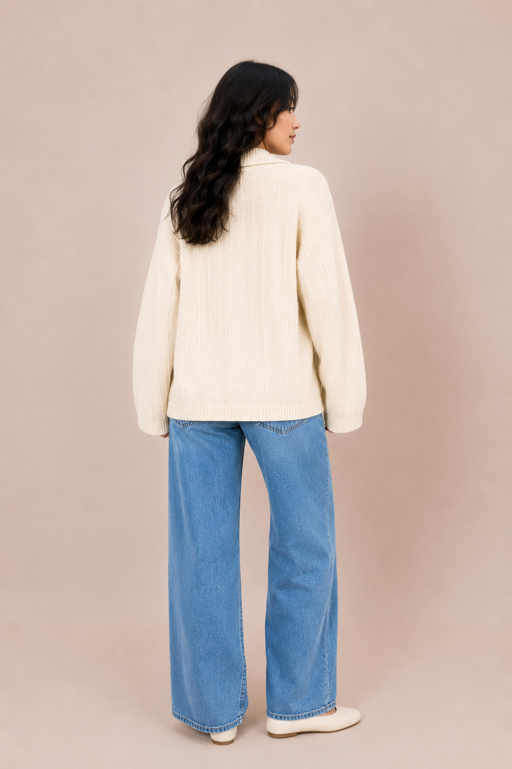 A woman is shown from behind wearing a cream ribbed knit sweater and wide-leg blue jeans against a solid beige studio background.
