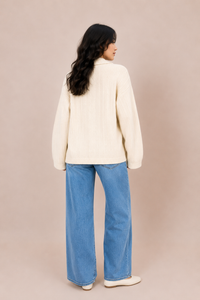 A woman is shown from behind wearing a cream ribbed knit sweater and wide-leg blue jeans against a solid beige studio background.
