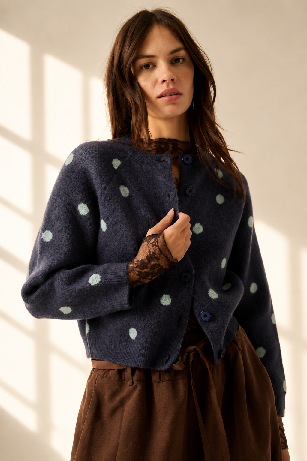 A woman wearing a navy blue knit cardigan with light green polka dots and brown pants, standing in a warmly lit room with soft window shadows.