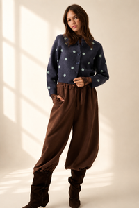 A woman modeling a dark navy blue knit cardigan with light blue-green polka dots, paired with brown wide-leg pants and dark brown boots, standing in a studio with warm window-light shadows.