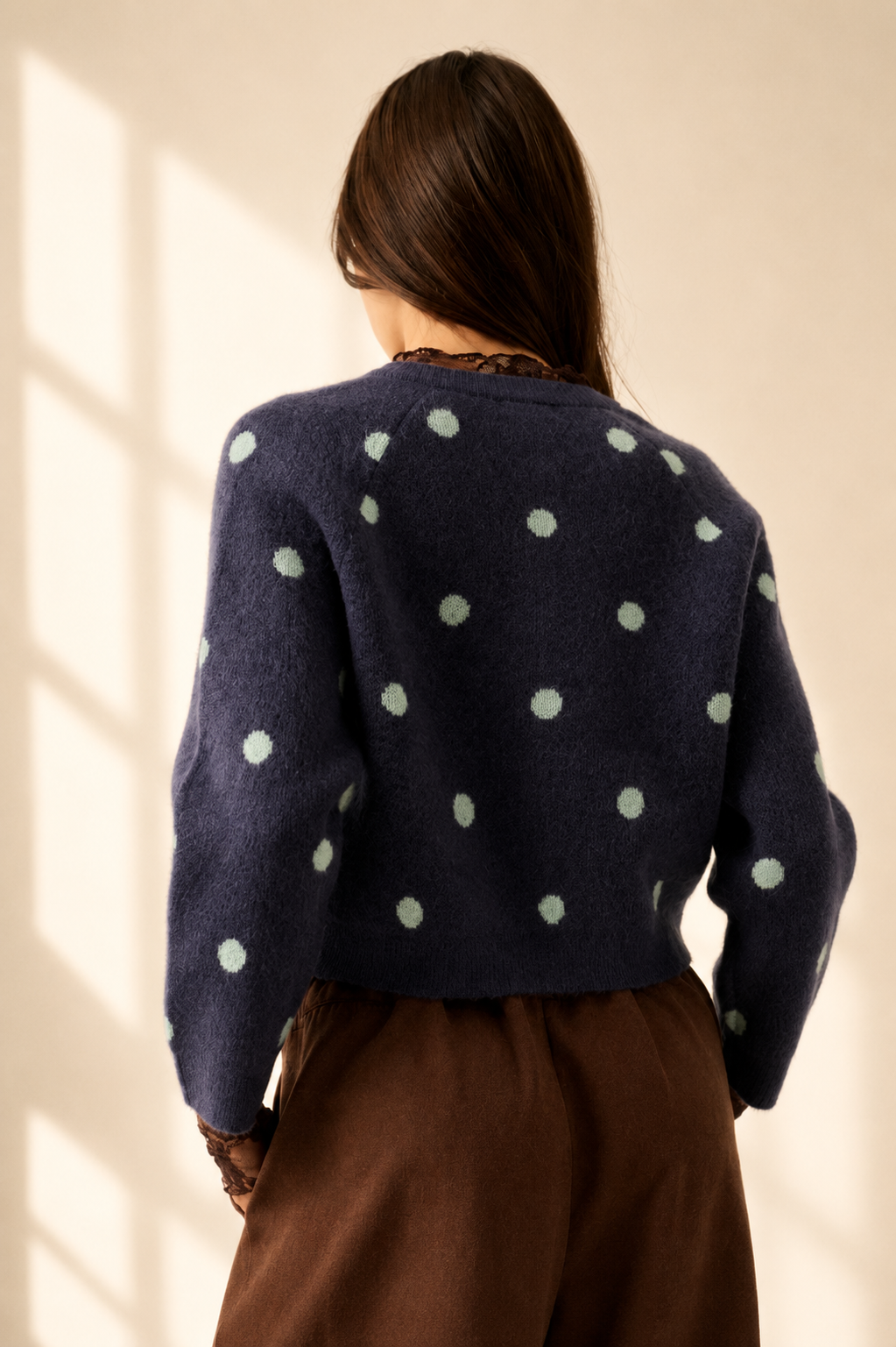 A woman viewed from behind wearing a navy blue fuzzy knit cardigan with light sage green polka dots, paired with brown trousers, set against a warm beige wall with soft window shadow light.