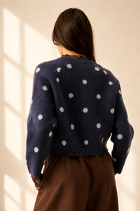 A woman viewed from behind wearing a navy blue fuzzy knit cardigan with light sage green polka dots, paired with brown trousers, set against a warm beige wall with soft window shadow light.