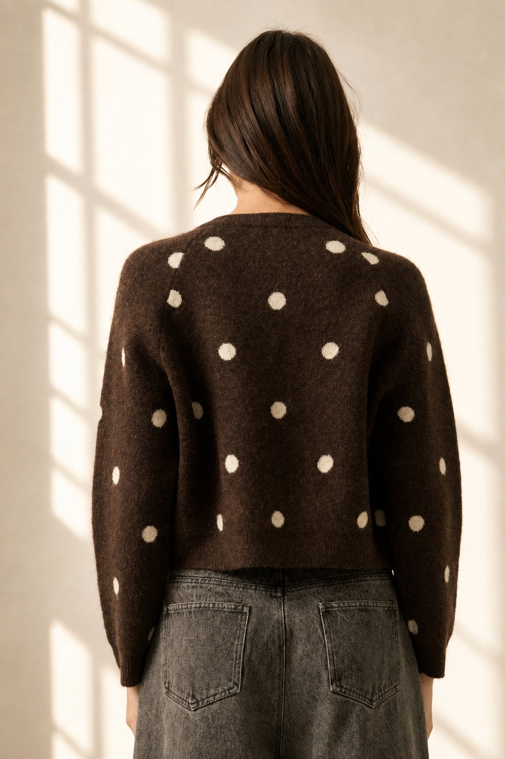 A woman viewed from behind wearing a dark brown knit sweater with large cream polka dots, paired with gray jeans, standing in a room with warm sunlight streaming through a window.