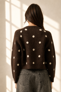 A woman viewed from behind wearing a dark brown knit sweater with large cream polka dots, paired with gray jeans, standing in a room with warm sunlight streaming through a window.