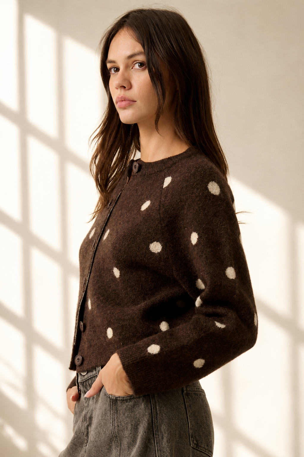 A woman wearing a dark brown fuzzy cardigan with cream-colored polka dots, paired with gray jeans, standing in a room with soft window light casting shadows on the wall.