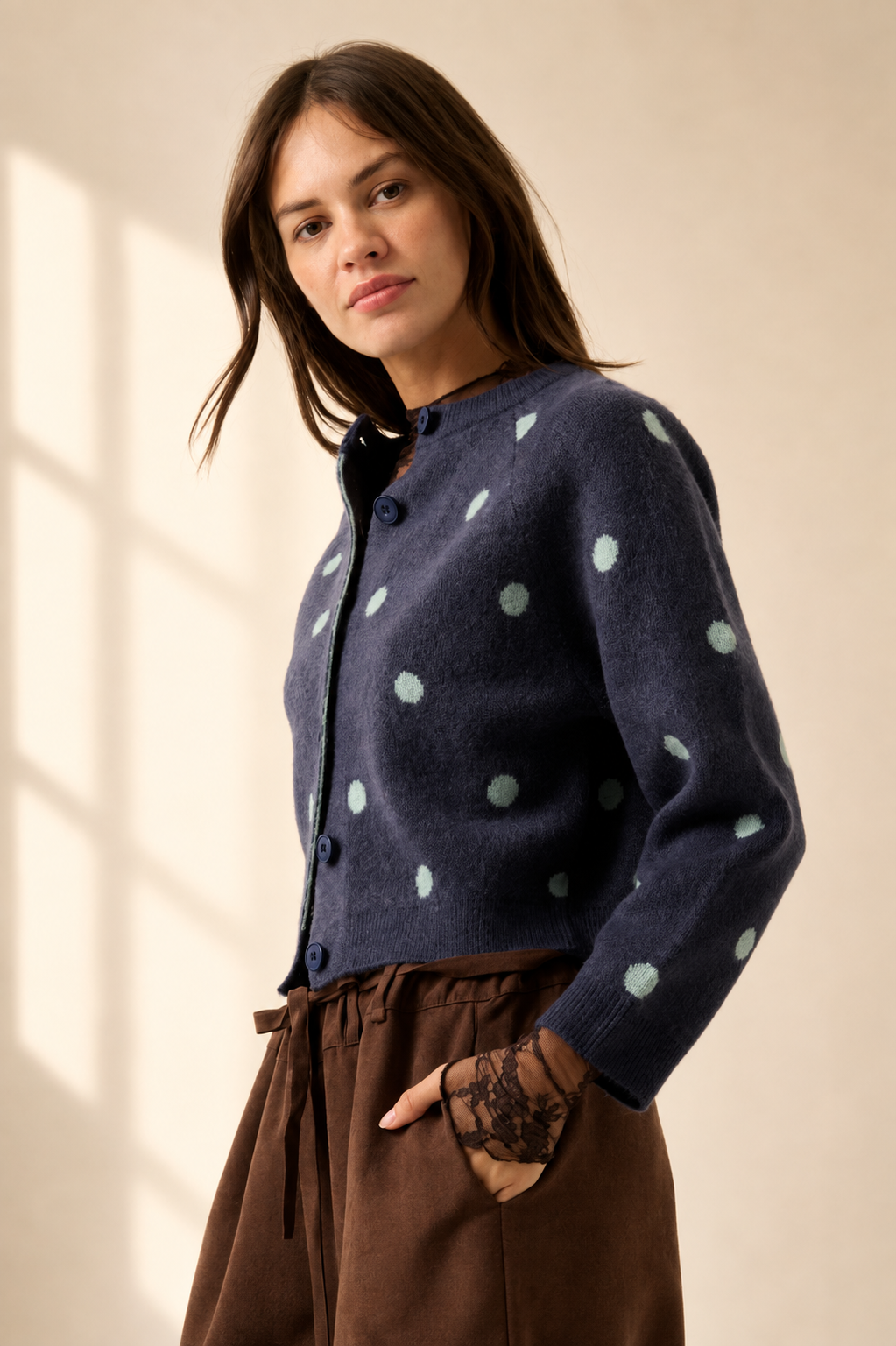 A woman wearing a navy blue knit cardigan with light green polka dots and brown wide-leg pants, standing in a softly lit room with window shadows.