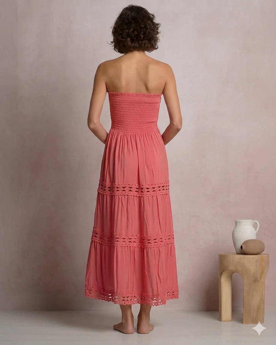 Back view of a woman wearing a coral pink strapless tiered maxi dress with a smocked bodice and crochet lace trim detailing, standing in a minimalist boutique setting.
