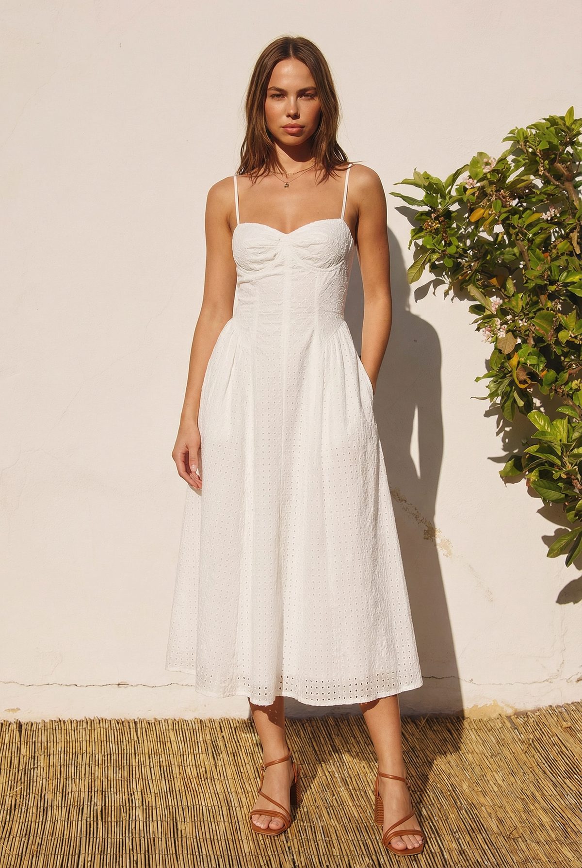 Woman wearing a white dress standing against a white wall with greenery.