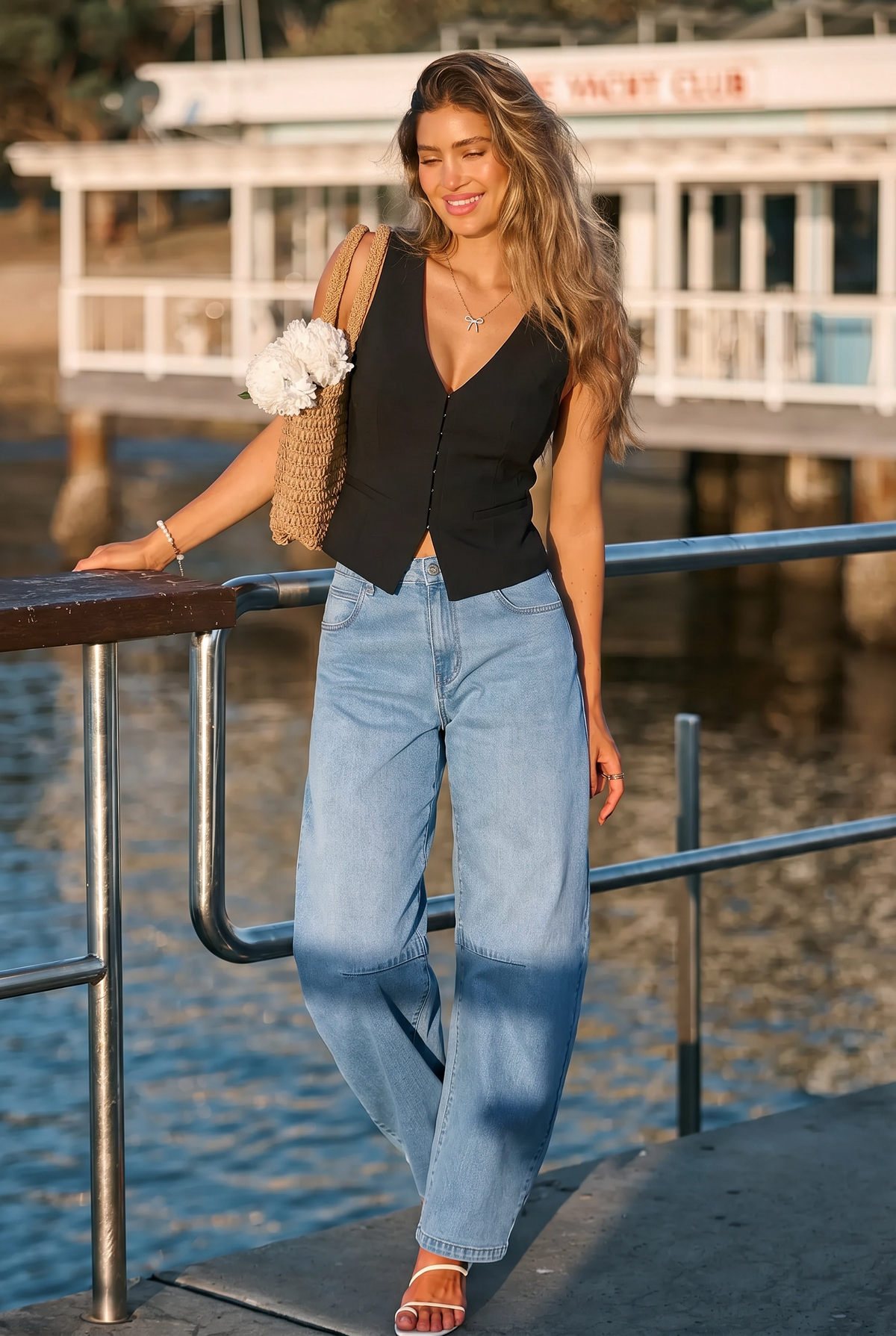 Woman standing by a waterfront wearing a black top and blue jeans, holding a straw bag.