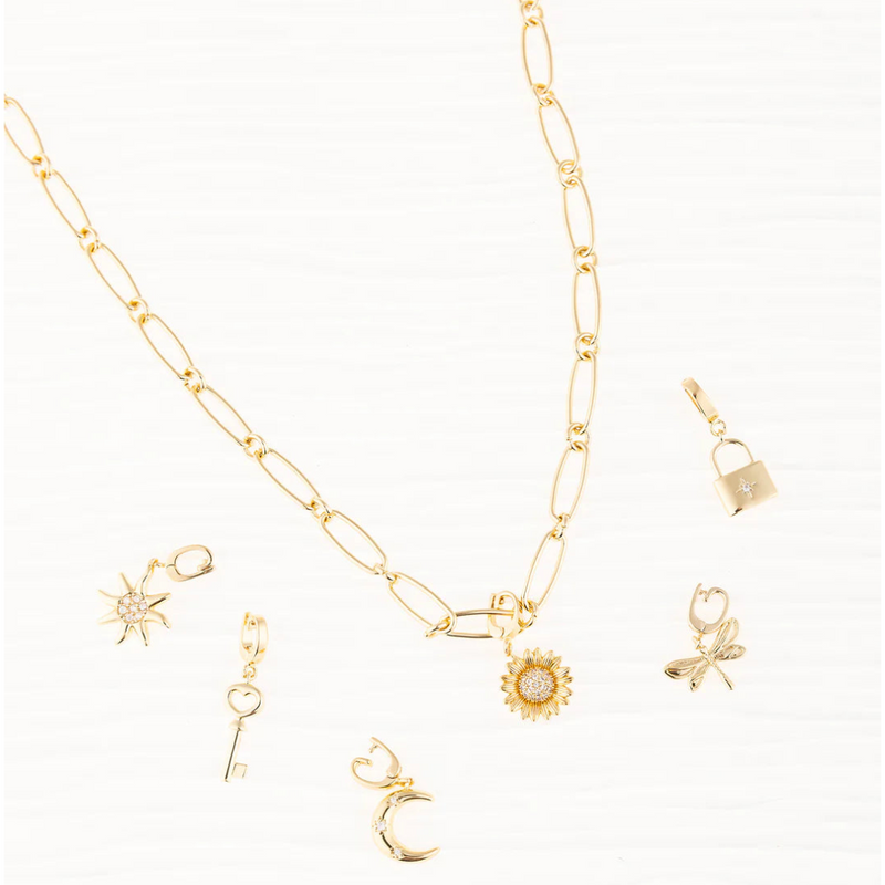 A gold paperclip chain necklace styled with various Michelle McDowell symbol charms, including a sun, key, moon, sunflower, dragonfly, and lock charm.