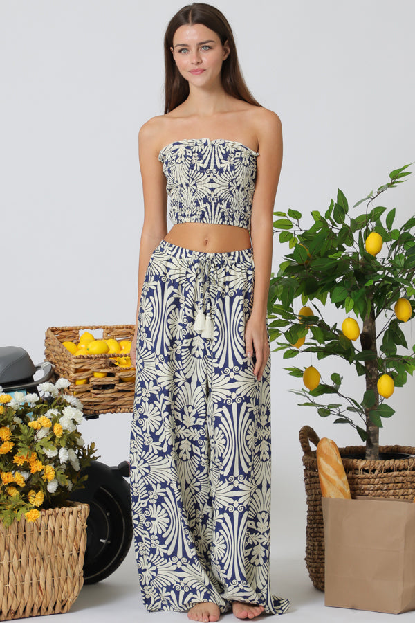 Woman wearing a strapless top and long pants with a floral pattern, standing in front of a lemon tree and baskets.