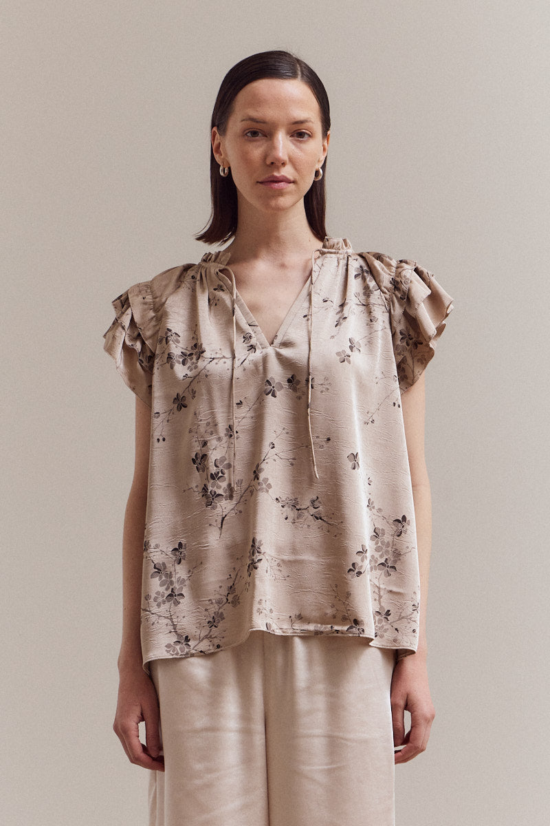 Model wearing the Perry Ruffle Sleeve Blouse in beige satin floral print — soft neutral blouse with flutter sleeves.