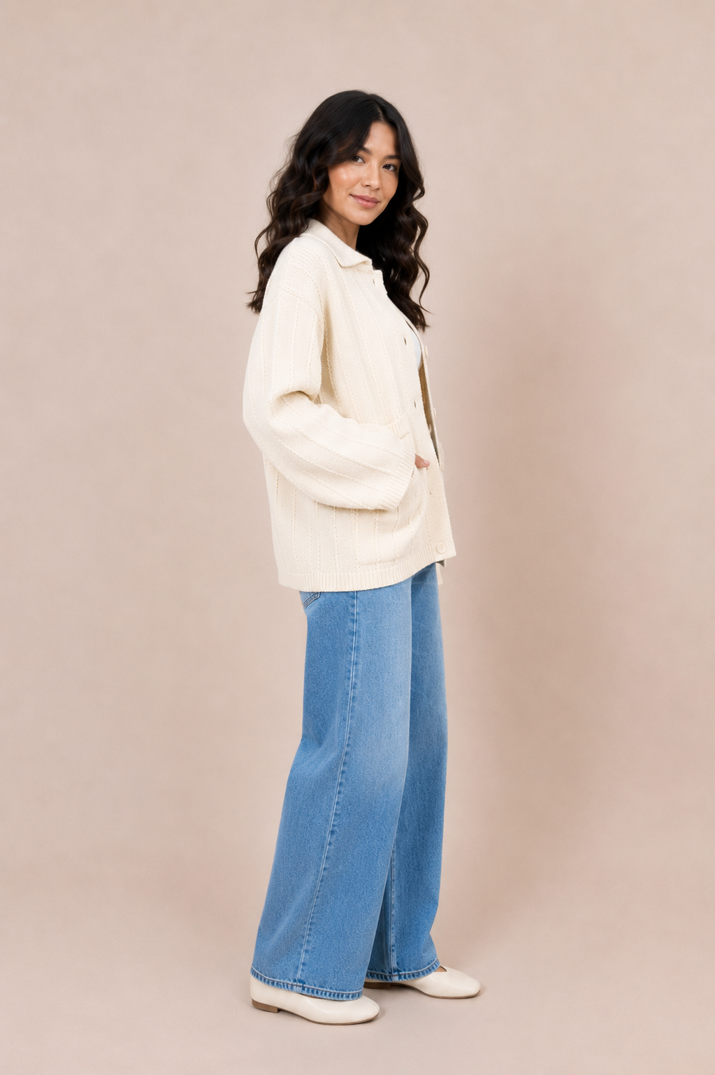 A woman modeling a cream-colored textured knit button-up shirt jacket and wide-leg blue jeans against a solid beige studio background.