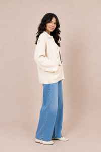 A woman modeling a cream-colored textured knit button-up shirt jacket and wide-leg blue jeans against a solid beige studio background.