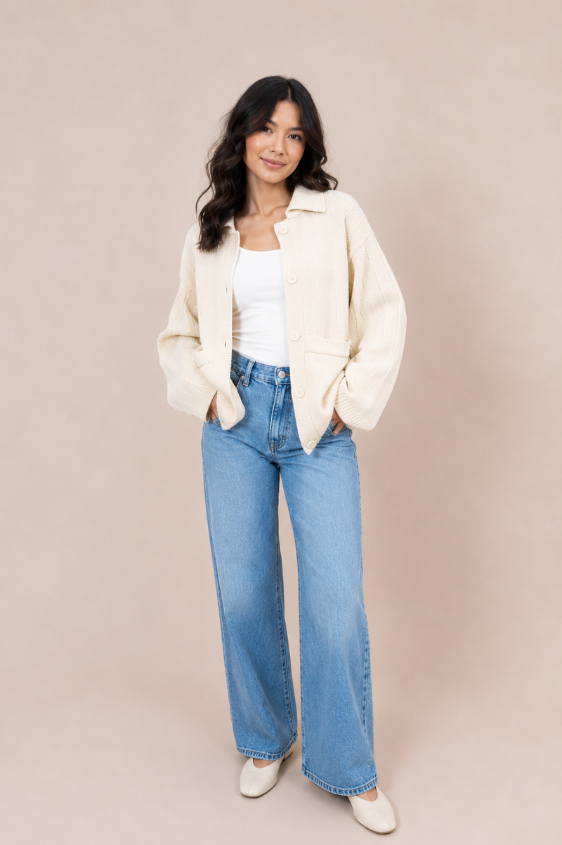 A woman modeling a cream-colored textured jacket over a white tank top and light blue wide-leg jeans, standing against a solid beige studio backdrop.