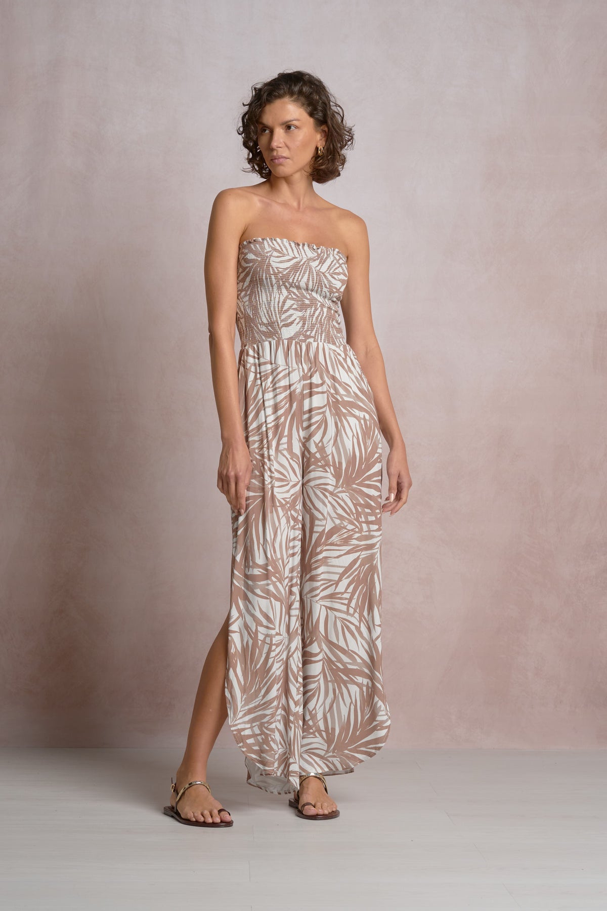 Front view of a model wearing the Isla strapless tropical print wide-leg jumpsuit featuring a smocked bodice and neutral Bali palm leaf pattern, styled with flat sandals for a resort wear look.