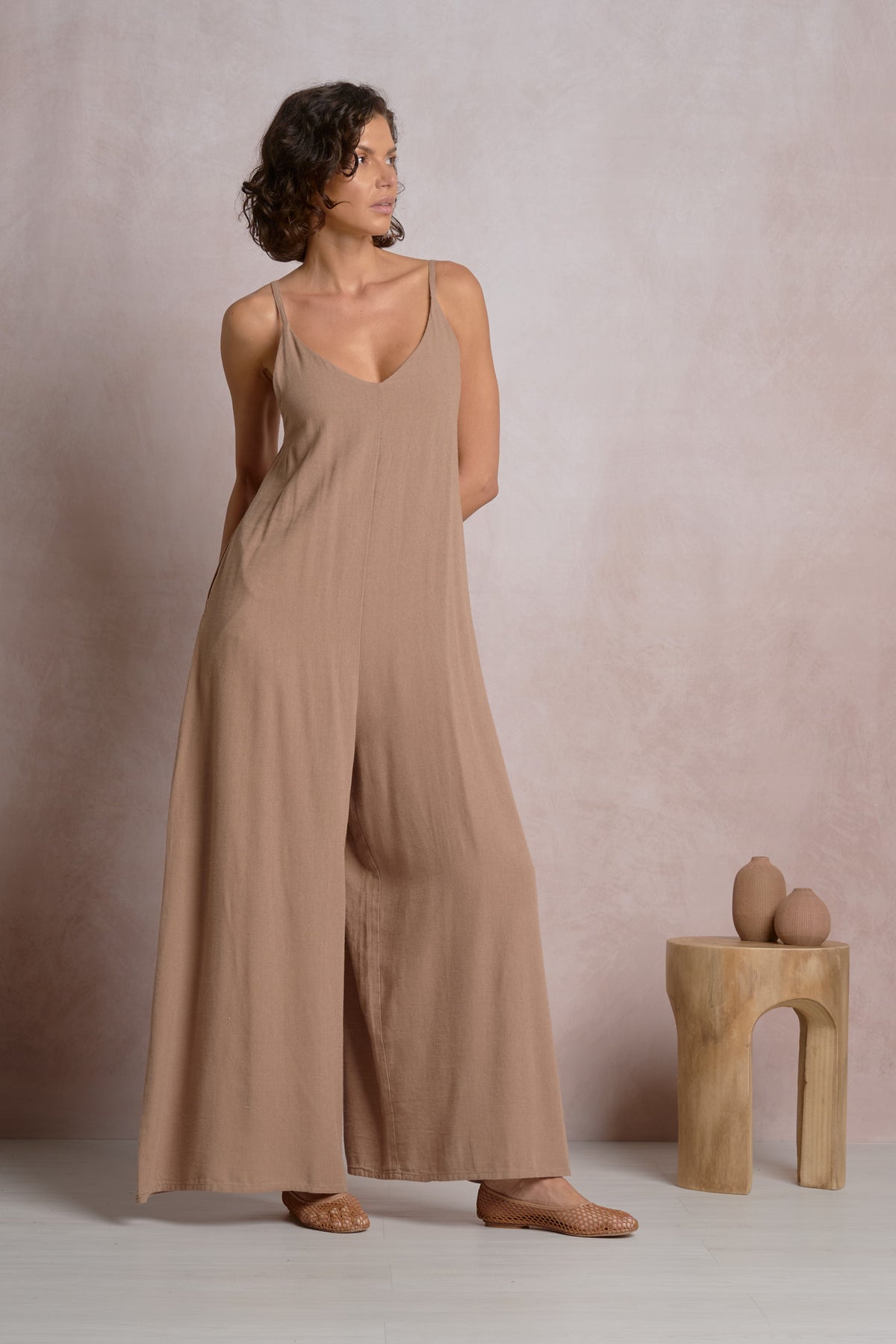 Front view of a model wearing the Courtney taupe spaghetti strap jumpsuit, featuring a V-neckline and dramatic wide-leg palazzo pants, styled with woven leather flats.