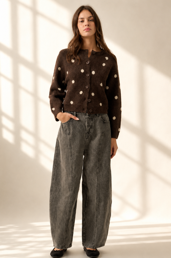 A woman stands in a studio wearing a brown button-front cardigan with white polka dots and wide-leg gray jeans, posed against a warm off-white wall with soft window-shadow light patterns.